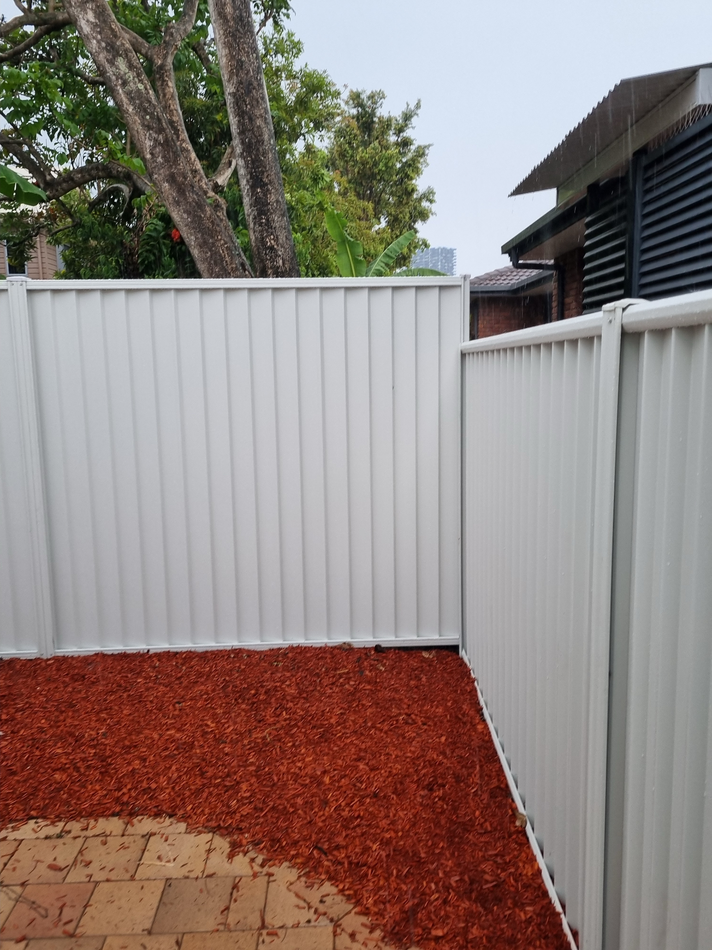 Cream Colorbond fence corner with mulched garden bed and pavers in foreground.